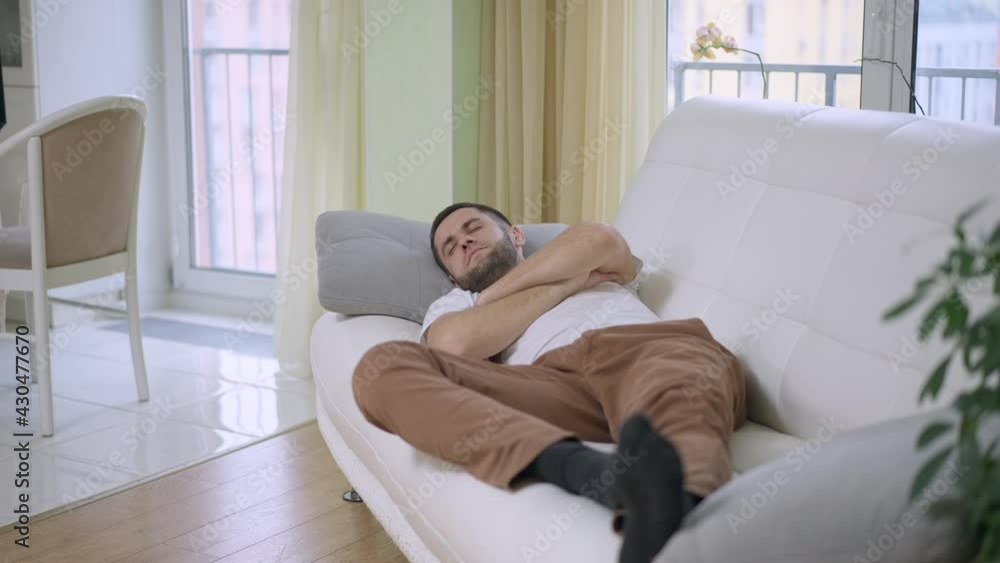 Tired young Caucasian man sleeping lying on couch in living room. Wide shot portrait of exhausted guy napping at home indoors. Lifestyle and tiredness concept