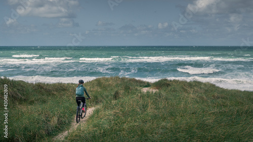 Woman with bike in sand dunes with grass at coast in Denmark, ocean in the background.
