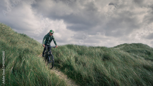 Man with bike in sand dunes with grass at coast in Denmark, dark clouds in dramatic sky.
