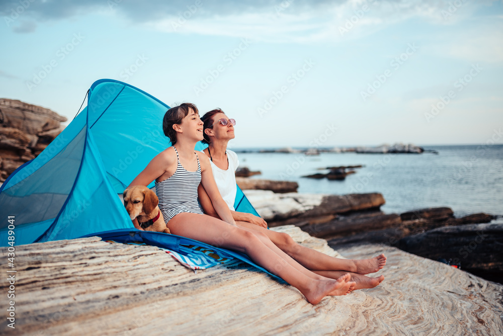 Mother and daughter looking into sunset while sitting in beach tent with dog