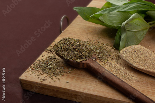 Wooden cutting board with vegetables or cooking ingredients; two wooden spoons with pepper and oregano and basil leaves; dark background