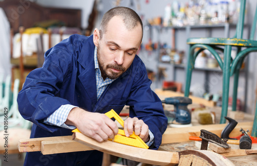 Confident man in uniform working in furniture repair workshop, measuring wooden plank before cutting