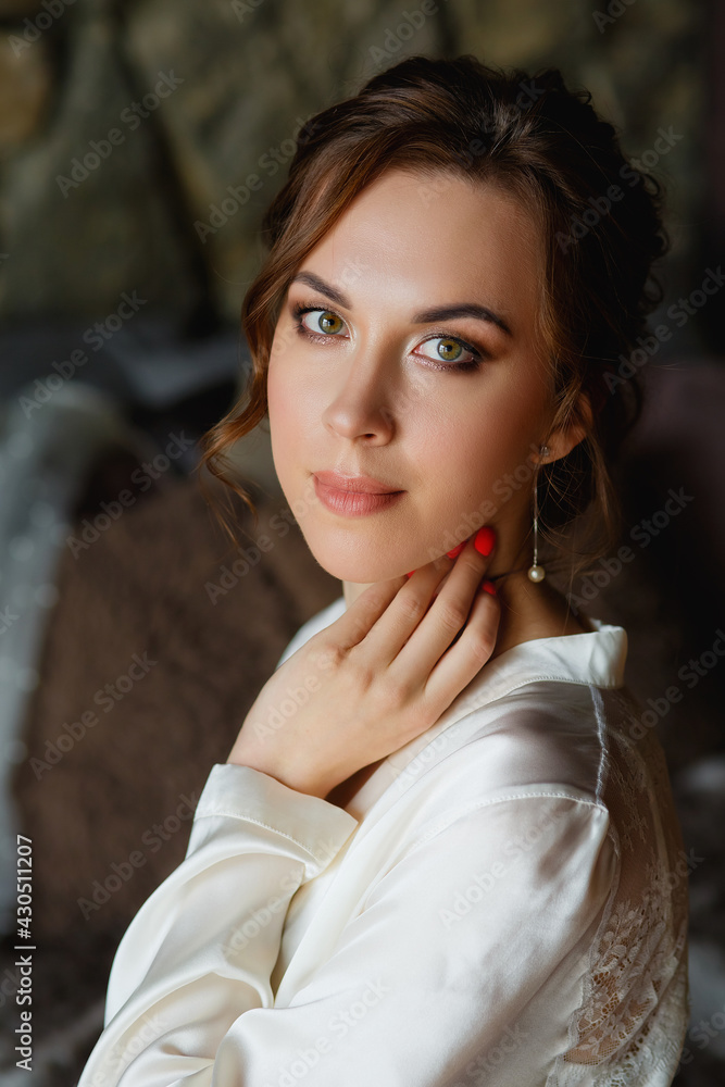Fototapeta premium Morning of the bride. A gorgeous bride, a beautiful woman in a white coat at the window. Morning of the bride before the wedding. Natural beauty and professional makeup.