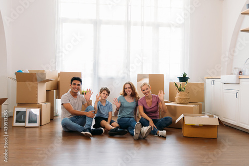 Cheerful caucasian parents and their cute son and daughter smiling and waving with hands on camera while sitting on floor at new apartment © twinsterphoto