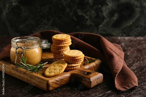 Board with tasty crackers and jar with peanut butter on dark background