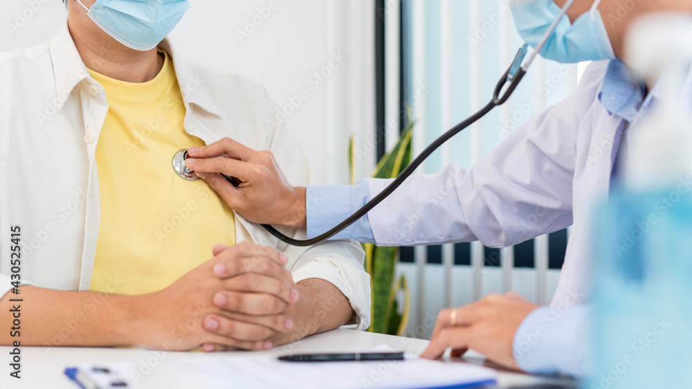 Medical concept a smart young male doctor wearing a mask using stethoscope to listen patient’s heart during examination in hospital room and wearing visor as preventing against Coronavirus outbreak 