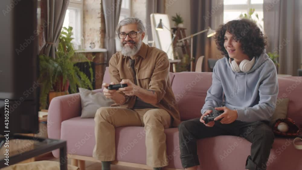 Happy mixed race teenage boy playing video game on console with middle aged father while sitting together on sofa at home Happy mixed race teenage boy playing video game on console with middle aged father while sitting together on sofa at home