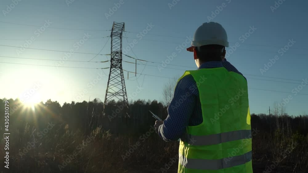 An energy engineer in special clothing checks the power line. Writes tasks to the tablet. Covers from the sun with his hand. Back view. An energy engineer in special clothing checks the power line. Writes tasks to the tablet. Covers from the sun with his hand. Back view.