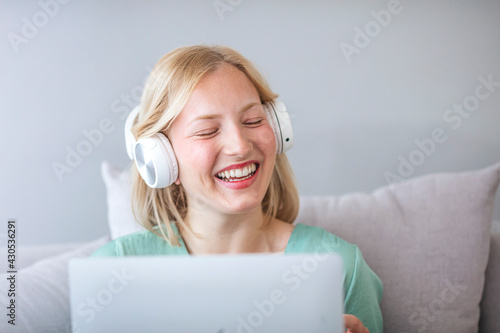 Smiling teenage female pupil sit on sofa in headphones wave hand to laptop screen greet teacher tutor at lesson consultation. Happy young woman say hi hello to friend speaking from home by video call