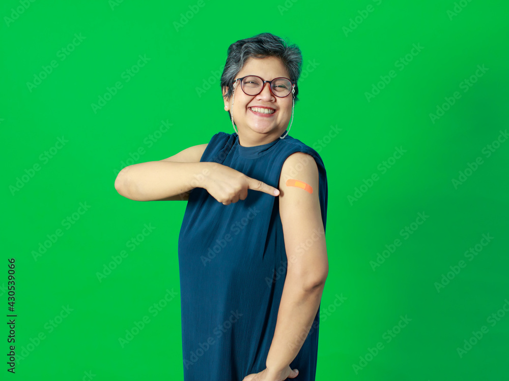 Fototapeta premium Adult senior Asian woman pointing and smile confident show shoulder with bandage plaster after getting Coronavirus of Covid-19 vaccine injection. Idea for vaccination of population against virus.