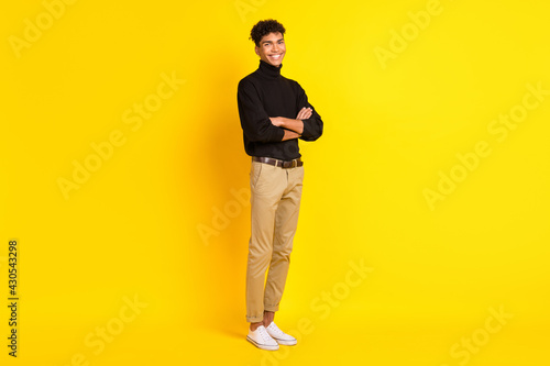 Full size profile photo of optimistic brunet guy stand crossed arms wear black sweater trousers sneakers isolated on yellow color background