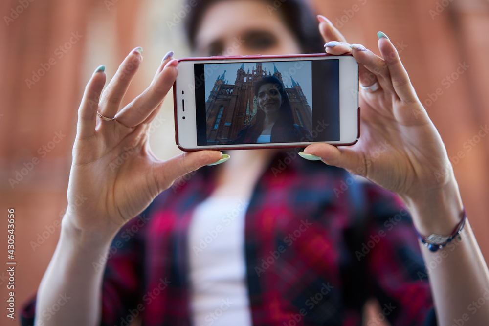 Blurred female tourist using modern mobile technology for clicking selfie images during travel sightseeing, millennial woman holding cellphone device with selective focus on touchscreen with photo