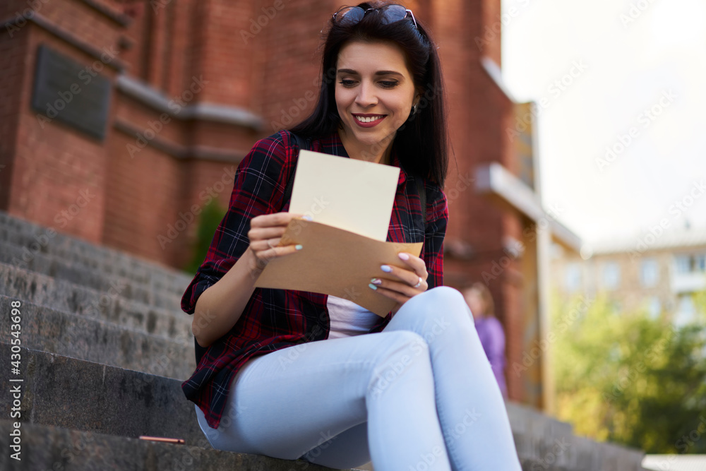 Fototapeta premium Happy millennial student with yellow envelope reading received mail notification during daytime in city, Caucasian hipster girl checking message and correspondence holding postal paper and postcard