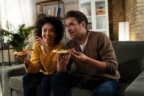  Cheerful young couple sitting on sofa at home. Happy woman and man eating pizza while watching a movie