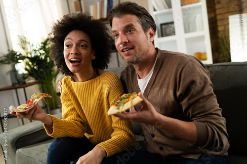 Cheerful young couple sitting on sofa at home. Happy woman and man eating pizza while watching a movie