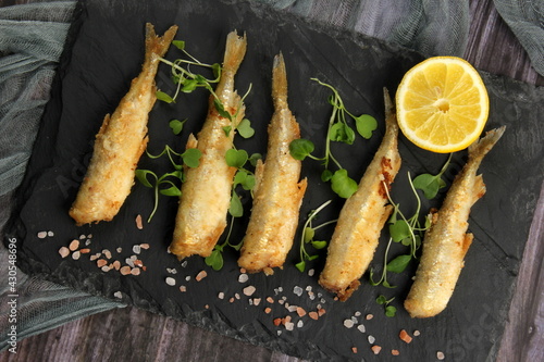 fried breaded fish served on a black stone, decorated with fresh lemon, dark wooden background. horizontal view from above
