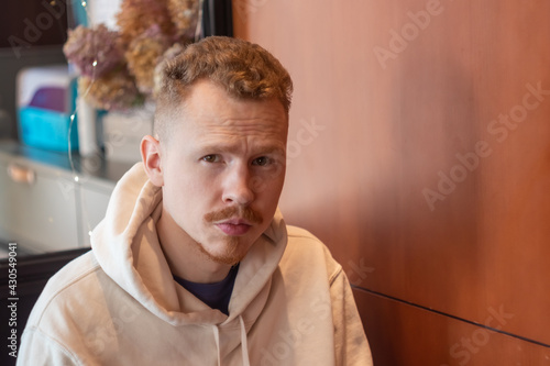 a man about 30 in a hoodie with a red mustache sits in a cafe