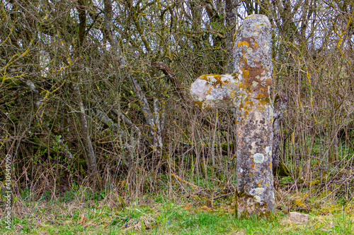 Old damaged stone atonement Cross covered with moss and lichen standing in front of a bush