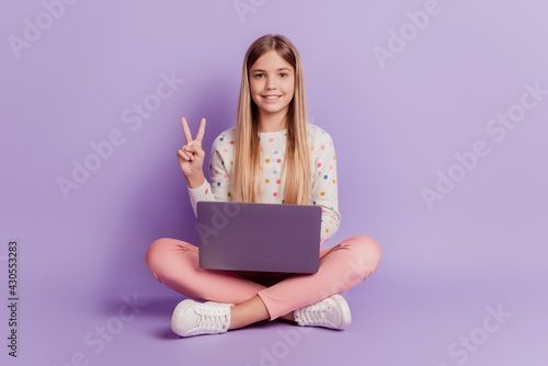 Positive kid sit legs crossed use laptop make v-sign isolated over violet background © Tetiana