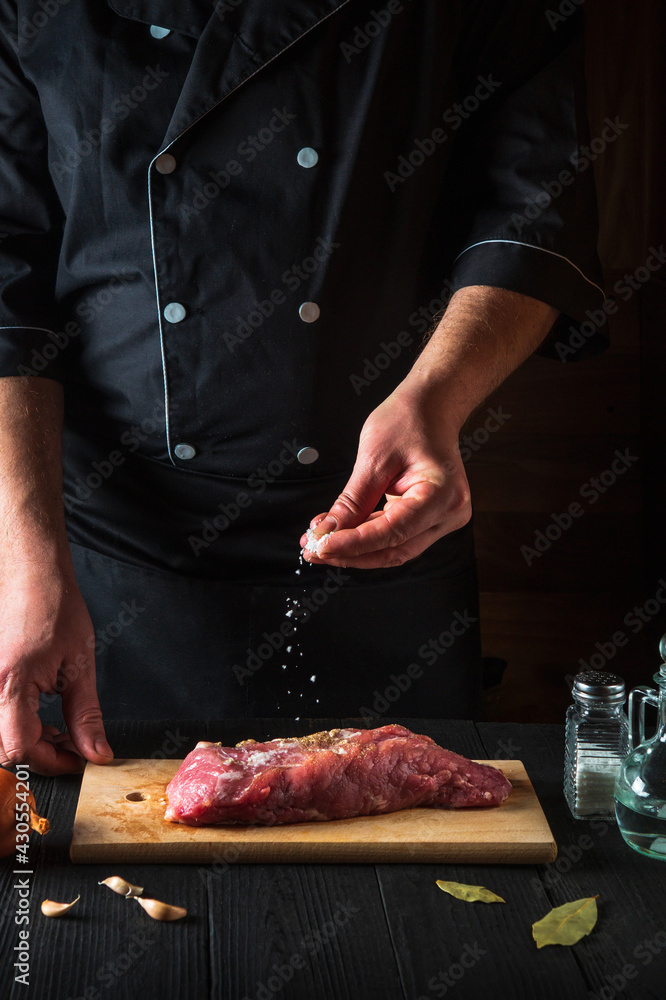 Chef sprinkles the raw beef meat with salt. Preparing meat before baking. Working environment in kitchen of restaurant or cafe