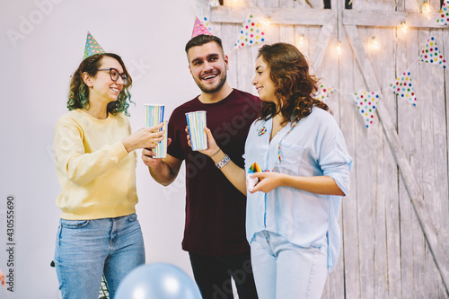 Positive male and female guests of birthday party in cone hats communicating holding cups with drinks enjoying celebration, smiling hipsters greeting best female friend clinking and wishing best