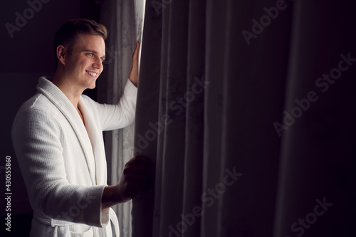 Man Wearing Bathrobe On Hotel Or Spa Break Opening Curtains