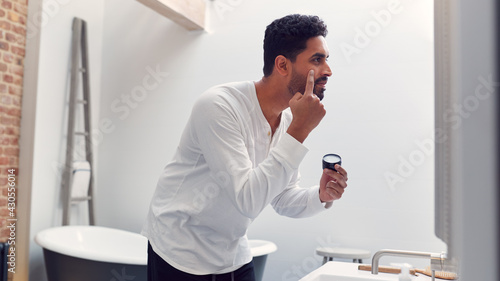 Man Wearing Pyjamas At Home In Modern Bathroom Putting On Moisturizer Looking In Mirror