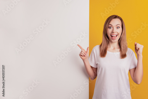 Young elegant woman pointing empty space white board raise fist on yellow wall
