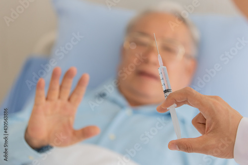 An old fat asian patient with gray hair wearing glasses and light blue shirt lying down in hospital bed hold his hand up try to stop doctor from inject vaccine to him because he afraid of the needle