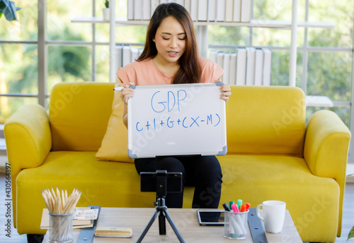 portrait of young asian woman sitting on a couch holding a signboard and marking pen teaching student online by using a smartphone at home. Learn from home concept