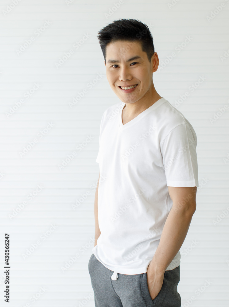 Medium shot portrait of a casual Asian handsome man with cute smiling in a white v-neck short sleeve t-shirt and gray pants with hands in pockets posing isolated with white background in the studio Medium shot portrait of a casual Asian handsome man with cute smiling in a white v-neck short sleeve t-shirt and gray pants with hands in pockets posing isolated with white background in the studio