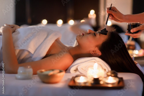 Masseurs hand do holding glass and brushing a facial mask herb black cream to on customer's face as sleep and covered warp by a white on her body