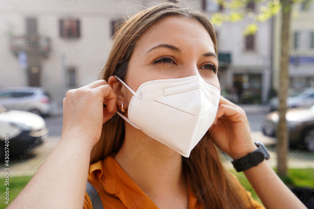 Close up of young woman wearing medical mask KN95 FFP2 in city street. Girl puts on protective face mask outdoors.