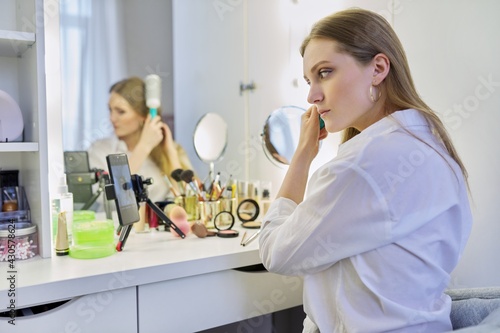 Young woman sitting at make-up table with mirror doing makeup and hairstyle