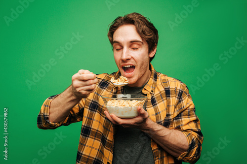 Young man eats flakes with milk and looks happy, isolated on green background