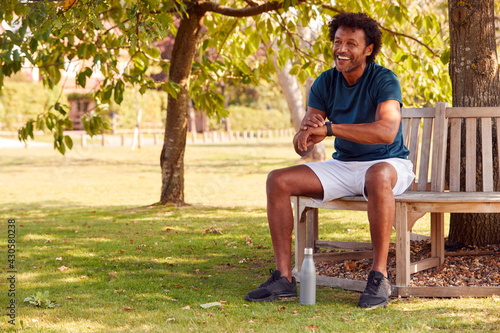 Man Wearing Fitness Clothing Sitting On Seat Under Tree Checking Activity Monitor On Smartwatch