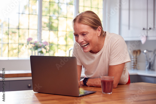 Mature Woman Wearing Fitness Clothing At Home Logging Activity From Smart Watch Onto Laptop