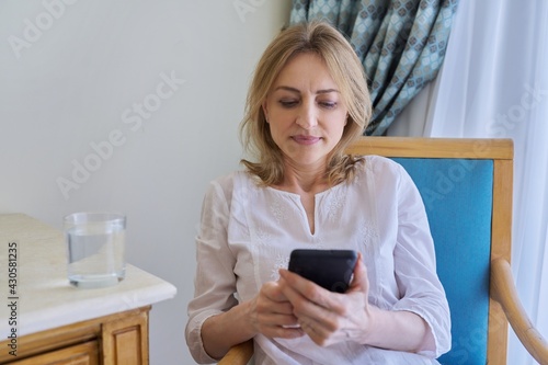 Serious middle aged woman with smartphone sitting on chair at home
