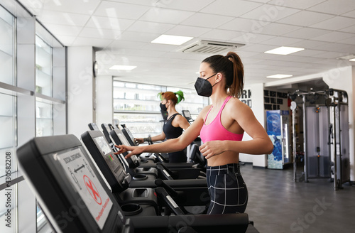 Two fit women exercising on treadmill wearing face mask in gym during pandemic