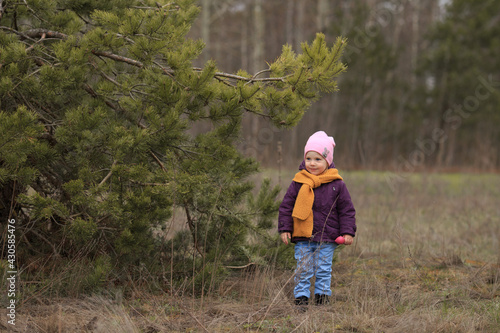 a girl in a pink hat and with a toy in her hand stands near a pine tree and looks away