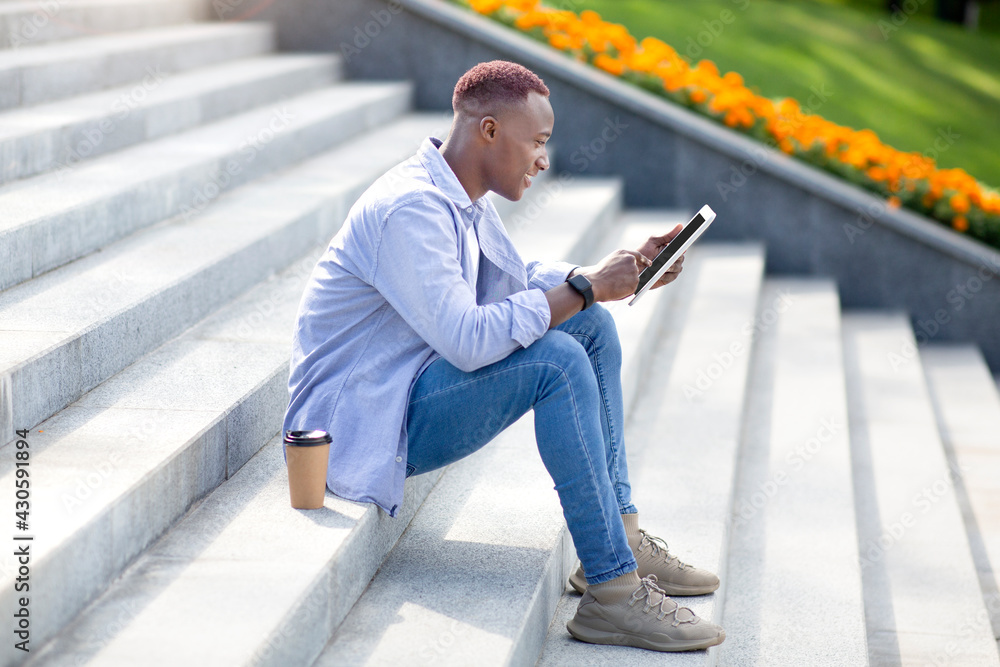 Side view of young black man with takeout coffee using tablet computer on stairs outdoors