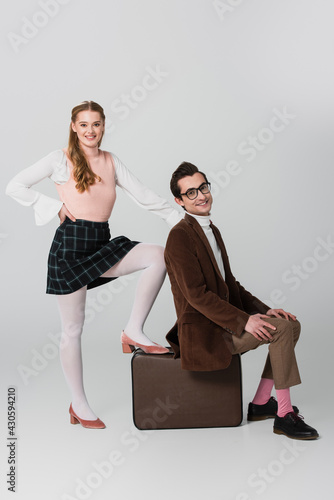 cheerful woman standing with hand on hip near boyfriend sitting on retro suitcase on grey background