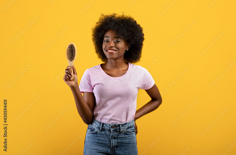 Happy african american woman holding wooden hair brush, smiling to camera over yellow studio background Happy african american woman holding wooden hair brush, smiling to camera over yellow studio background
