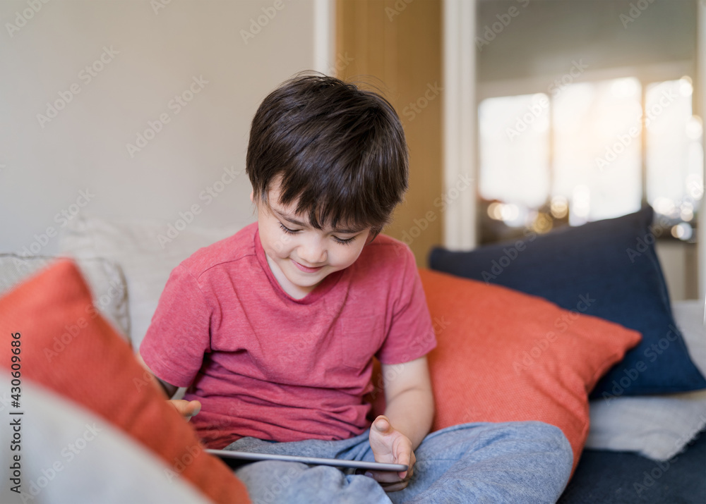 Authentic Kid sitting on sofa watching cartoons or playing games on tablet,Child boy using digital pad learning lesson on internet,Home schooling,Distance learning online education concept Authentic Kid sitting on sofa watching cartoons or playing games on tablet,Child boy using digital pad learning lesson on internet,Home schooling,Distance learning online education concept