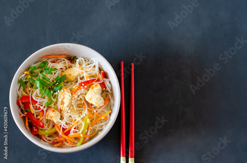Asian salad with glass noodles, chicken and vegetables - carrots, bell peppers and sesame seeds on a gray plate with chopsticks. Flat lay. Horizontal orientation. Copy space