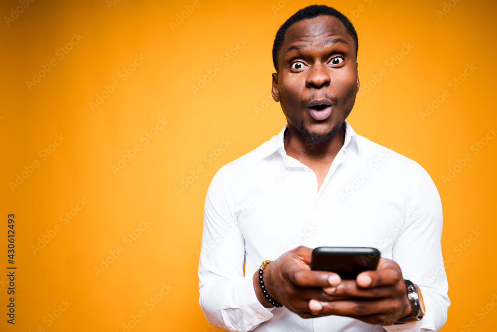 Handsome african american man holding a phone in his hand and is surprised by its power while standing on yellow background Handsome african american man holding a phone in his hand and is surprised by its power while standing on yellow background