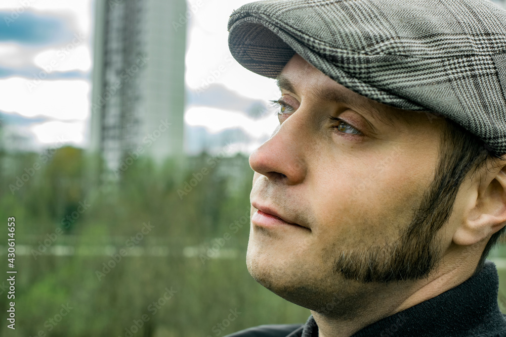 A man with sideburns and a cap looks thoughtfully into the distance