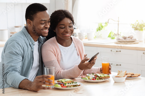 Millennial Black Man And Woman Having Breakfast And Using Smartphone In Kitchen
