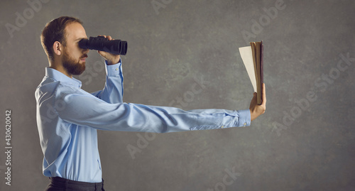 Profile view of serious young guy who needs glasses looking in binoculars and reading book he's holding in long extended arm. Humorous concept of dealing with presbyopia, bad eyesight, vision problems