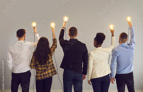 Group of young multiethnic business professionals holding up shining Edison light bulbs standing on gray studio background, back view. Creative innovative thinking, people developing own idea concept